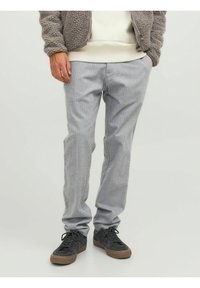 Uomo che indossa pantaloni grigi chiari, sneakers nere con suole marroni e una giacca in pile beige, con le mani in tasca.