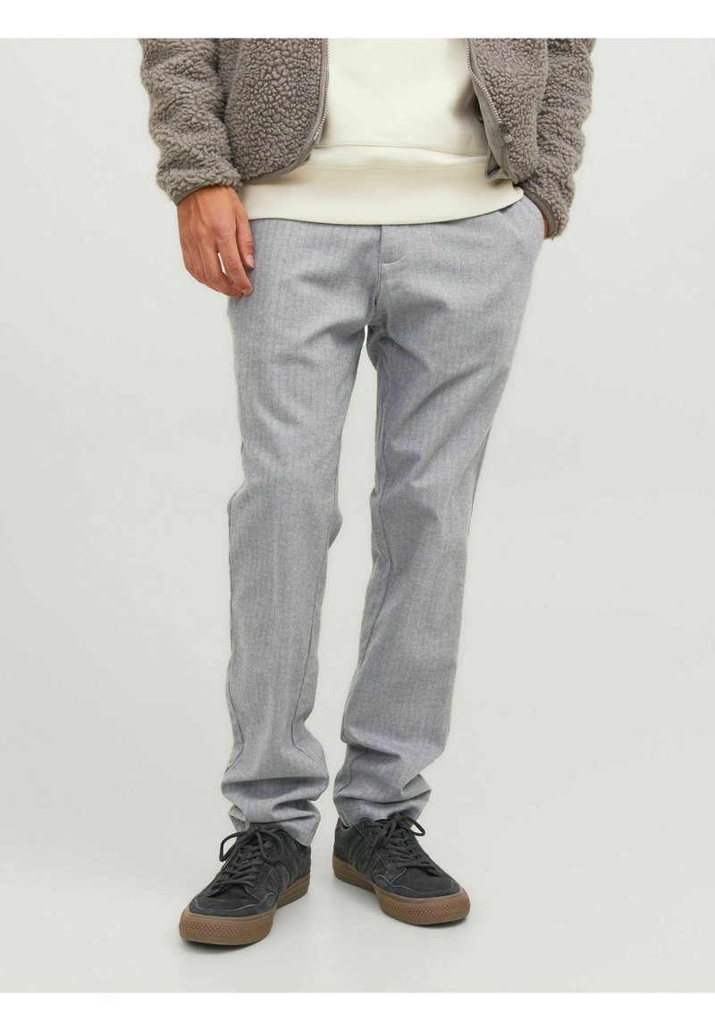 Uomo che indossa pantaloni grigi chiari, sneakers nere con suole marroni e una giacca in pile beige, con le mani in tasca.