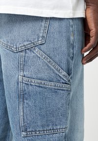 Un pantalon cargo en denim bleu clair présente plusieurs poches angulaires, des coutures visibles et une coupe décontractée. Le tissu a une texture légèrement usée.