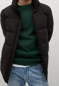 Schwarze Steppjacke mit Reißverschluss, die gesteppte Abschnitte aufweist. Darüber trägt die Person einen grünen Pullover und kombiniert ihn mit hellblauen Jeans.