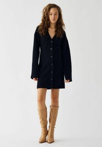 Jūras spēku zilas adītas kleitas ar V veida kakla izgriezumu, garām piedurknēm un priekšējiem pogām. Nēsāta ar brūnām augstiem zābakiem līdz ceļiem. Gluda tekstūra, ikdienišķs dizains.