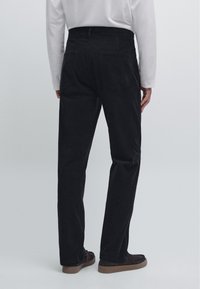 Svarta manchesterbyxor med rak skärning, featuring två bakfickor och en slät textur. Bärs med en vit långärmad skjorta.