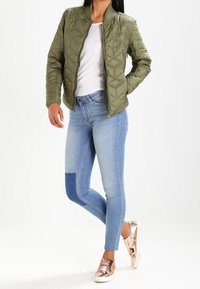 Quilted olive green bomber jacket, white t-shirt underneath, light blue skinny jeans with a dark patch, and rose gold slip-on sneakers.