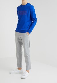 Felpa blu con logo rosso, pantaloni tuta grigi a gamba affusolata e sneakers bianche. I materiali sembrano morbidi con una texture liscia.