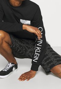 Czarna koszula z długim rękawem z białym logo "CALVIN KLEIN" na rękawie i kieszeni; połączona z czarnymi szortami z powtarzającym się wzorem logo; czarne sneakersy.