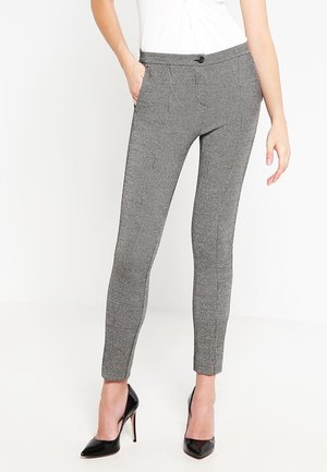 Kobieta ubrana w szare, dopasowane spodnie w jodełkę z guzikowym pasem, w połączeniu z czarnymi szpilkami i białą bluzką, stojąca.