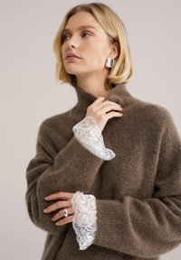 Pull en maille marron avec un col montant. Présente une bordure en dentelle blanche aux poignets. La texture semble douce et confortable, avec une coupe décontractée.