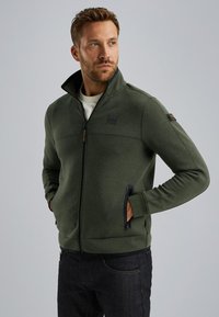 Tmavozelená bundu na zips s vysokým golierom, bočnými vreckami a textúrou pripomínajúcou mäkký fleece. Má logo na hrudi a kontrastné zipsy.