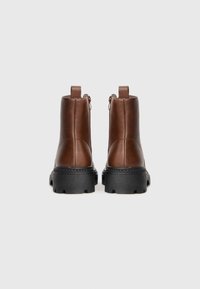 Par de botas de cuero marrón hasta el tobillo con suelas negras gruesas, mostradas desde la parte trasera sobre un fondo gris claro liso.