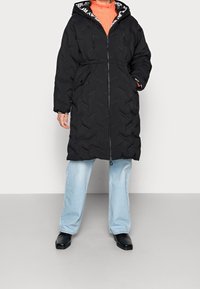 Černý zateplený kabát s utaženým pasem, plným zipem a kapucí, která má kontrastní potisk. Oblečené s světle modrými širokými kalhotami a černými botami.