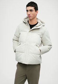 Casaco puffer leve em tecido off-white suave, com capuz, fecho de correr à frente e textura acolchoada diagonal. Inclui bolsos com aba.