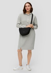 Sivá mikinová šaty s kapucňou a dlh�ými rukávmi, so bočnými rozparokmi, nosené s bielymi teniskami. Čierna crossbody taška s hladkým povrchom.
