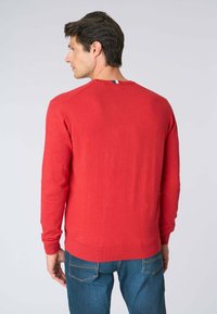 Maglione rosso a maglia con scollatura a girocollo, polsini e orlo a coste. Presenta un piccolo dettaglio blu e bianco sul retro della scollatura. Indossato con jeans blu.