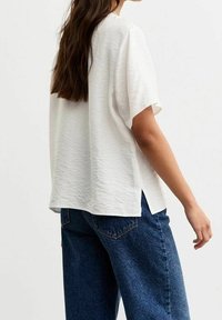 Bílý top s krátkými rukávy a texturovaným povrchem, s volnějším střihem a bočními rozparky, kombinovaný s modrými, vysokým pasem džínami.