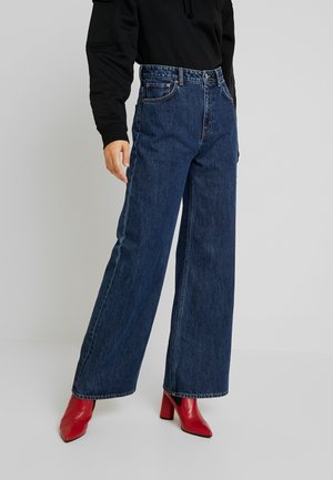 Persoon met donkerblauwe wijde jeans en rode enkellaarsjes met hak, met een zwart shirt met lange mouwen tegen een effen achtergrond.