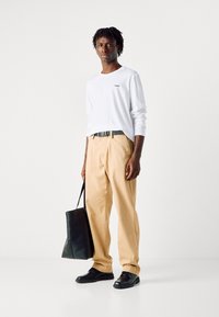 Cămașă albă din bumbac cu mâneci lungi cu logo-ul "HUGO", pantaloni bej, pantofi negri din piele și o geantă tote neagră într-un decor minimalist.