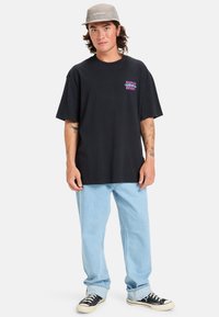 Camiseta negra de algodón con un ajuste amplio, con un gráfico colorido en el pecho. Combinada con jeans de color azul claro y zapatillas negras. Gorra gris.
