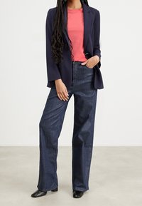 Kvinna står med långa flätor, klädd i en marinblå kavaj, korallfärgad t-shirt, vida mörk denimjeans och svarta skor.