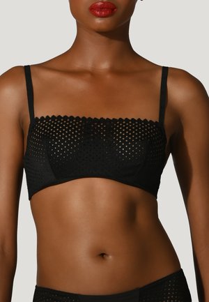 Frau trägt einen schwarzen perforierten Bandeau-BH mit dünnen Trägern, dargestellt von den Schultern bis zur Mitte des Oberkörpers vor einem einfarbigen hellen Hintergrund.