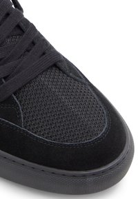 Zapatilla negra que presenta una combinación de malla texturizada y materiales de ante. Punta redondeada con cordones planos y suela de goma.