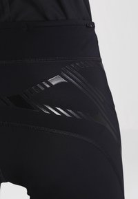 Close-up van zwarte sportleggings met glanzende geometrische patronen op het bovenbeen en heupgebied.