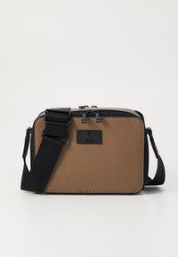 Hnedá textúrovaná kožená crossbody taška s čiernymi detailami, nastaviteľným popruhom a zipsovým uzáverom. Obsahuje logo značky na prednej strane.