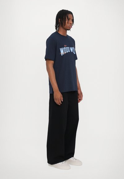 Camiseta de algodón azul marino con gráfico de "WOOD WOOD" en azul y rojo. Combinada con pantalones anchos negros y zapatillas blancas. Diseño sencillo.
