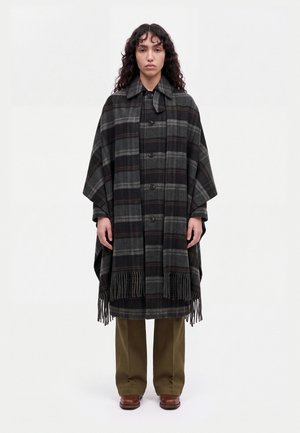 Un mannequin se tient de face, vêtu d’un manteau-cape à carreaux gris foncé avec des franges, d’un pantalon olive et de chaussures marron, sur un fond blanc uni.
