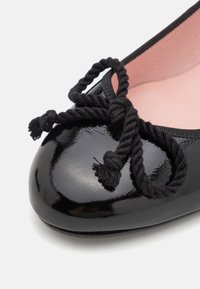 Ballerine en cuir verni noir avec un bout arrondi, ornée d'un nœud décoratif en cordon noir torsadé à l'avant. Doublure intérieure rose.
