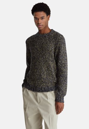 Hombre con cabello oscuro y rizado, viste un suéter de punto negro y gris y pantalones color crema, de pie con las manos en los bolsillos.