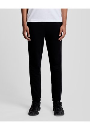 Person trägt schwarze schmal geschnittene Hose und schwarze Sportschuhe, steht mit entspannten Händen seitlich am Körper vor einem einfarbigen Hintergrund.