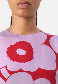 Close-up van het onderste deel van het gezicht en de nek van een persoon die een lavendelkleurige top draagt met grote rode abstracte bloemmotieven.