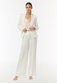 Biały żakiet w stylu dopasowanym na beżowej satynowej bluzce, w połączeniu z luźnymi białymi spodniami i białymi sandałami na paski. Gładka faktura, minimalistyczny design.