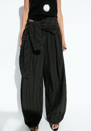 Femme portant un pantalon noir à jambes larges à rayures avec un détail de nœud surdimensionné à la taille et des sandales noires à bout ouvert.