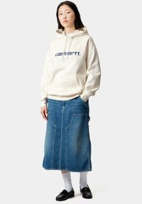 Sudadera crema con logo azul marino, combinada con una falda de mezclilla larga y azul que presenta dos grandes bolsillos frontales y un corte recto. Zapatos negros y calcetines blancos.