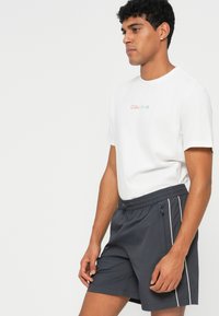 White cotton t-shirt with multicolored "Calvin Klein" logo, paired with dark gray swim shorts featuring a drawstring and side stripes.