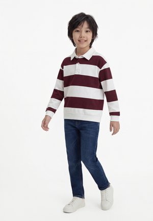 Gutt står og smiler, iført en langermet polo-skjorte med burgunder- og hvite striper, blå jeans og hvite joggesko.