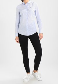 Kvinde iført en lyseblå skjorte med knapper, sorte stramme bukser og hvide sneakers, stående foran en ensfarvet baggrund.