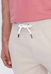 Pantalons de jogging beiges avec taille élastique, dotés d'un cordon de serrage blanc avec embouts en métal noir. Texture de tissu lisse, design basique.