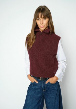 Woman with straight brown hair wears a maroon sleeveless sweater over a white shirt and blue jeans, hands in pockets, facing forward.