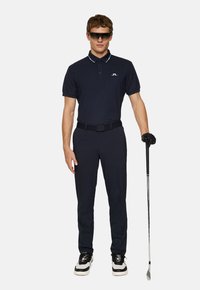 Marinblå polo skjorta med vita krageaccenter, kombinerad med matchande byxor. Bärs med svarta handskar och solglasögon, håller en golfklubba.