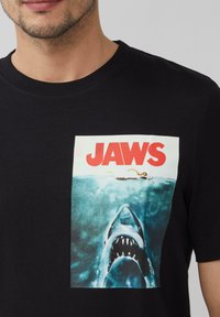 Zwart katoenen T-shirt met een grafische print van een haai en een zwemmer, met "JAWS" in vetrode letters boven het beeld.