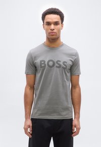 Νέος άνδρας με κοντά σγουρά μαλλιά φορώντας γκρι μπλουζάκι με τη λέξη "BOSS" τυπωμένη με μεγάλα γράμματα, στέκεται μπροστά σε απλό λευκό φόντο.