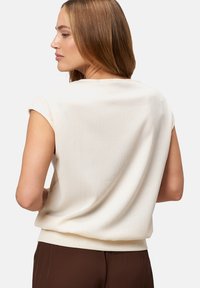 Cream short-sleeve top featuring a textured fabric, a relaxed fit, and a ribbed hem, paired with brown trousers in the background.