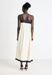 Lång krämfärgad klänning med svarta detaljer, med tunna axelremmar, en samlad ryggdesign och en solid kant vid fållen. Bärs med svarta klacksandaler.