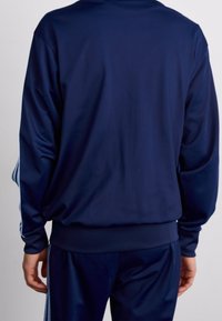 Mörkblå träningsjacka, tillverkad av slätt tyg, har långa ärmar och en ribbad nederkant. Sidostråkar i en ljusare nyans är synliga.