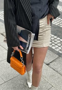 Blazer a righe nere sopra una camicia blu navy, mini gonna beige arricciata, borsa arancione trapuntata con catena argentata e stivali bianchi alla caviglia.