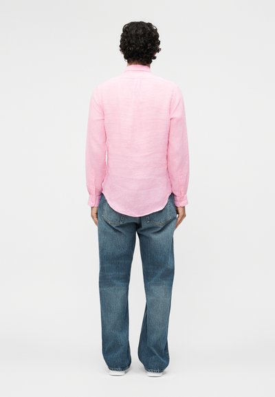 Persona ar cirtainiem tumšiem matiem, valkājot garu rozā kreklu un vaļīgas zilas džinsa bikses, stāv ar muguru pret vienkāršu fona virsmu.