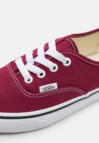 Gros plan d'une basket Vans en toile grenat avec des lacets blancs, des coutures blanches et une semelle en caoutchouc blanche avec une bande noire.