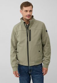 Veste softshell khaki avec un col haut, fermeture éclair à l'avant, accents noirs et deux poches latérales, associée à un jean bleu. Texture lisse.